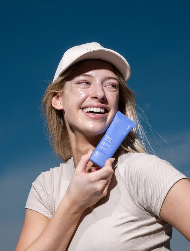 Woman wearing Sun & Dusted Cap Jeuneora Logo Cap and posing with tube of sunscreen