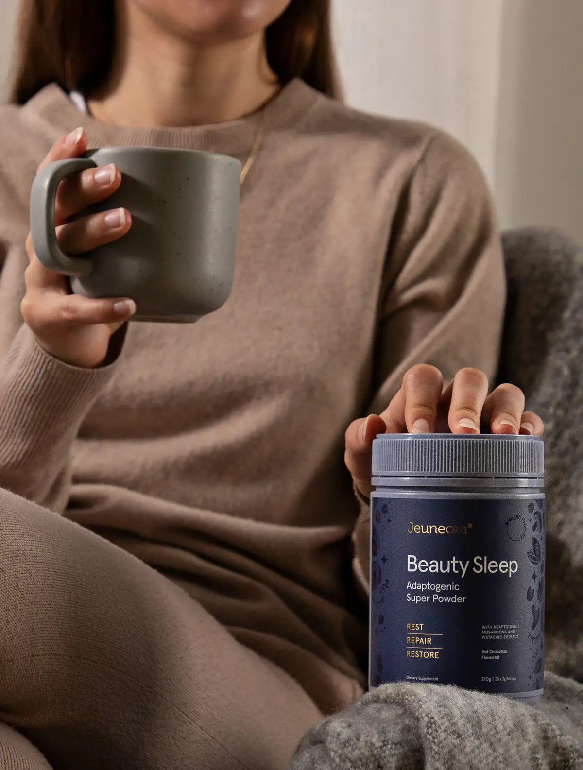 A woman sits in a chair and holds a container of Beauty Sleep sleep supplement and a mug