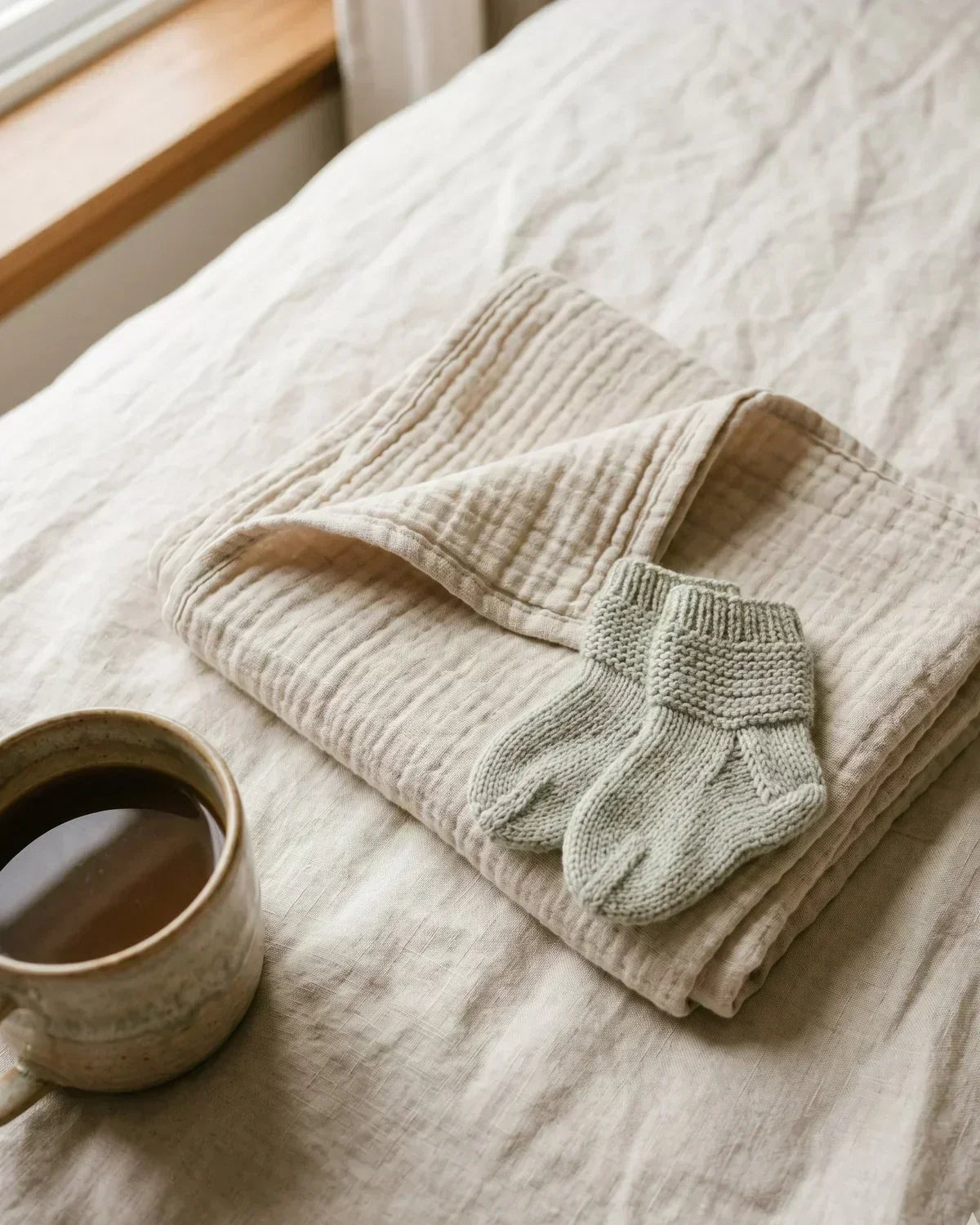 A folded cream muslin baby blanket with small sage green knitted baby socks resting on top, beside a ceramic mug of coffee on soft linen bedding by a sunlit window.