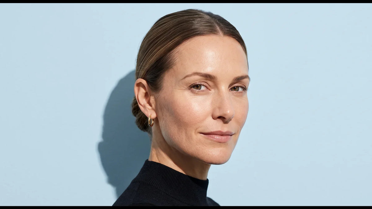 Close portrait of a woman in her forties with natural skin texture, hair pulled back, against a pale blue wall