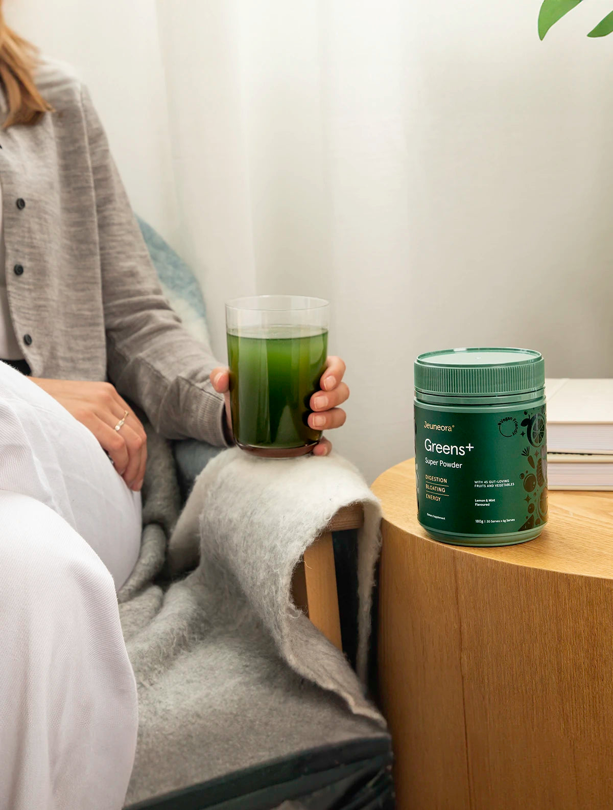 A woman sits on a chair holding a glass of Jeuneora Greens+ Super Powder. Next to her there is a container of the supplement on a side table.
